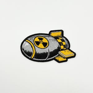May include: Embroidered patch depicting a bomb design. The patch features a grey body with yellow detailing, including a radiation symbol and a chequered pattern. The patch is outlined in black.