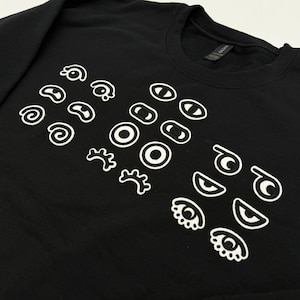 May include: A black long-sleeve sweatshirt featuring a variety of white embroidered eye designs. The graphic consists of multiple pairs of eyes in different shapes, including spirals, circles, and lashes, arranged across the front of the garment.