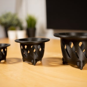 Peut inclure: Trois pots de fleurs en plastique noir avec un motif en treillis et trois pieds. Les pots sont disposés par ordre de taille, le plus grand pot au premier plan et le plus petit pot à l'arrière-plan.