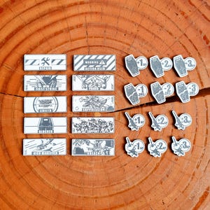 Puede incluir: Un conjunto de fichas y tarjetas rectangulares blancas y grises para un juego, con iconos y texto como "REPAIR", "FIRST STRIKE" y "BLOCKER". Las fichas presentan números y símbolos, lo que sugiere un juego de estrategia.