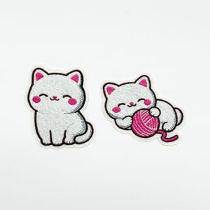 May include: Two embroidered patches of cartoon cats. One cat sits, the other plays with a ball of pink yarn. Both cats have white fur, pink cheeks, and black outlines. Ideal for clothing or accessories.