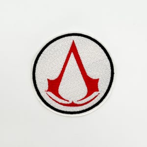 Può includere: Toppa rotonda ricamata con il logo di Assassin's Creed. La toppa ha uno sfondo bianco e un bordo nero. Il logo è una forma a 'A' rossa stilizzata.