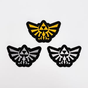 May include: Three embroidered patches featuring the Hylian Crest from The Legend of Zelda. One patch has a yellow and black design, while the other two have a black and white design. The patches are on a white background.