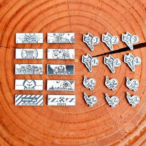May include: A collection of game tokens and markers with a sci-fi theme. Rectangular tokens feature text like "FIRST STRIKE" and "BLOCKER." Circular tokens display numbers and symbols. The items are arranged on a wooden surface.