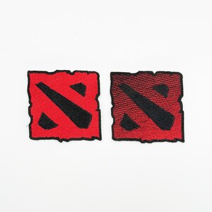 May include: Two embroidered patches featuring the Dota 2 logo. The patches are square with a jagged edge. One patch is solid red with a black diagonal slash and a black triangle. The other patch has a red to black gradient.