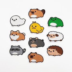 Puede incluir: Colección de parches bordados con varios animales de dibujos animados, incluyendo un castor, una foca, una rana, una oveja, un perro, un oso, un mapache, un erizo y una criatura amarilla. Cada parche tiene un borde blanco.