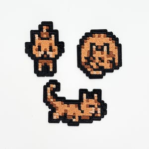 以下が含まれることがあります： さまざまなポーズの茶色の猫を描いた、3つのピクセルアートパッチ。1つのパッチは立っている姿、別のパッチは猫の顔、そして最後のパッチは横たわっている猫を示しています。各パッチには黒い縁があり、白い背景にあります。