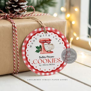 Pode incluir: Uma etiqueta de presente redonda com um padrão xadrez vermelho e branco, com o texto "MADE WITH LOVE". A etiqueta apresenta um gráfico de misturador vermelho e as palavras "Butter Pecan COOKIES". Um pinha e barbante completam o visual festivo.