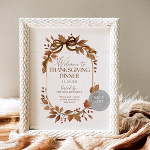 May include: A framed Thanksgiving dinner sign with a white textured frame. The sign features a brown floral wreath and text that reads "Welcome to Thanksgiving Dinner" and "hosted by: The Miller Family."