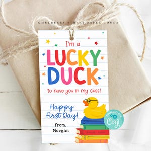 Könnte beinhalten: Ein Geschenkanhänger mit dem Text "I'm a LUCKY DUCK to have you in my class!" in bunten Buchstaben. Eine gelbe Ente mit Brille sitzt auf einem Bücherstapel. Der Anhänger ist mit einer Schnur an einem Geschenk befestigt.