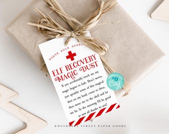Puede incluir: Una etiqueta de regalo blanca con rayas rojas y blancas en la parte inferior. La etiqueta tiene una cruz roja y el texto "ELF RECOVERY MAGIC DUST" en rojo. El texto dice: "Si me tocas accidentalmente, mi magia comienza a desvanecerse. No te preocupes, solo espolvorea un poco de este polvo mágico en mi cabeza, cuenta hasta tres, luego muéveme a la estantería y déjame estar. Por la mañana estaré como nuevo, todo gracias a ti!"