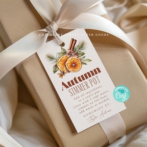 Może przedstawiać: Biała etykieta prezentowa z akwarelową ilustracją pomarańczy, cynamonowych patyczków i zieleni. Tekst na etykiecie brzmi "Autumn Simmer Pot" i zawiera instrukcje do przygotowania simmer pot. Etykieta jest przywiązana beżową wstążką do brązowego pudełka prezentowego.
