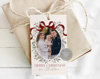 Peut inclure: Une étiquette cadeau de Noël avec une photo de famille dans un cadre ovale, ornée d'un ruban rouge et de verdure festive. L'étiquette indique "Merry Christmas from The Olsens" en script bordeaux. L'étiquette est attachée à un cadeau emballé.