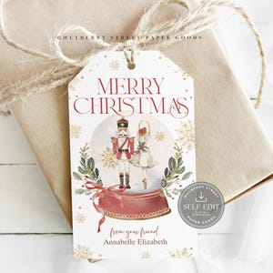 以下が含まれることがあります： 「MERRY CHRISTMAS」の赤い文字が入ったクリスマスギフトタグ。くるみ割り人形とバレリーナのイラスト、雪の結晶と緑が描かれています。タグは、ひもで包まれたギフトに結び付けられています。