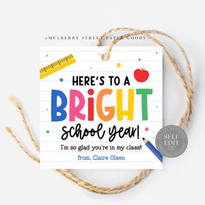 May include: A square gift tag with a school theme. The tag has the text "Here's to a Bright School Year!" in colorful letters, with a red apple and a yellow ruler graphic. The tag is tied with a natural twine.