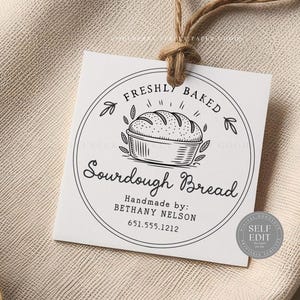 Puede incluir: Una etiqueta cuadrada blanca con un diseño circular que presenta un pan de masa madre en una cesta, con las palabras "Freshly Baked Sourdough Bread". La etiqueta está atada con hilo y incluye el nombre y número de teléfono del fabricante.