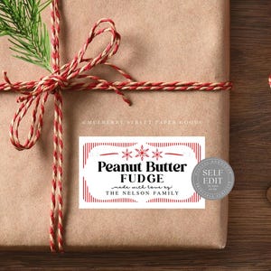 Può includere: Un regalo avvolto con un fiocco di spago rosso e bianco e un rametto di sempreverde. Un'etichetta rettangolare recita "Peanut Butter Fudge" con accenti di fiocchi di neve e "made with love by The Nelson Family."