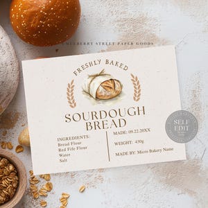 Op de afbeelding: Een label voor zuurdesembrood met de tekst "FRESHLY BAKED SOURDOUGH BREAD". Het label bevat ingrediënten, gewicht (430g) en een aquarel illustratie van brood. Het label is omgeven door brood en granen.