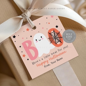 May include: A gift tag with a Halloween theme. The tag is light pink with a white ghost, a black spiderweb, and the word "BOO" in pink. The text "Here's a little treat for you! Happy Halloween" is also on the tag. The tag is tied to a gift with a cream ribbon.