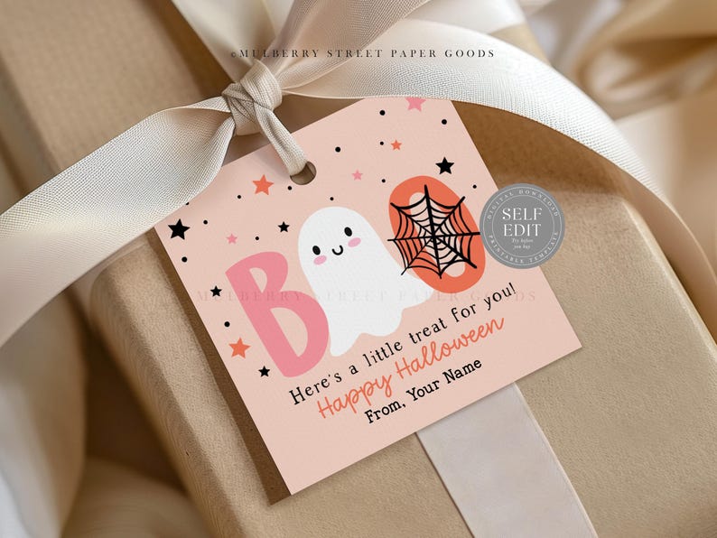 May include: A gift tag with a Halloween theme. The tag is light pink with a white ghost, a black spiderweb, and the word "BOO" in pink. The text "Here's a little treat for you! Happy Halloween" is also on the tag. The tag is tied to a gift with a cream ribbon.