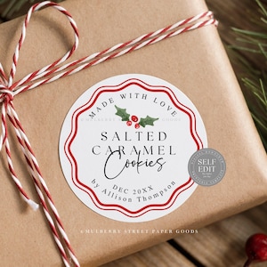 Pode incluir: Um presente embrulhado em papel castanho com um rótulo redondo que diz "Made with Love, Salted Caramel Cookies". O rótulo tem uma borda vermelha e detalhes de azevinho. O presente é amarrado com barbante vermelho e branco.
