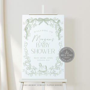 Pu&ograve; includere: Un cartello bianco per baby shower su un cavalletto, con un design floreale verde e la scritta "Benvenuti al baby shower di Morgan". Il cartello include la data e il luogo. Un adesivo "Self Edit" &egrave; a destra.