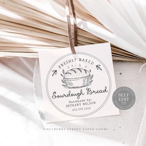Puede incluir: Una etiqueta cuadrada con una cinta marr&oacute;n presenta un dise&ntilde;o circular con las palabras "FRESHLY BAKED Sourdough Bread". El dise&ntilde;o incluye un pan en una cesta y el texto "Handmade by: BETHANY NELSON 651.555.1212."
