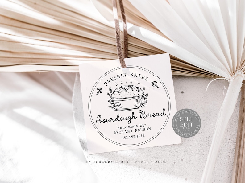 Puede incluir: Una etiqueta cuadrada con una cinta marr&oacute;n presenta un dise&ntilde;o circular con las palabras "FRESHLY BAKED Sourdough Bread". El dise&ntilde;o incluye un pan en una cesta y el texto "Handmade by: BETHANY NELSON 651.555.1212."