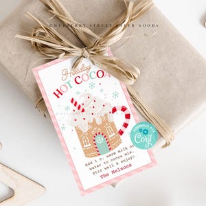 以下が含まれることがあります： 「Holiday HOT COCOA」と書かれたタグが付いた、包装されたギフト。タグには、ホイップクリームとキャンディケーンが付いたジンジャーブレッドハウスのマグカップが描かれています。タグには、指示と「The Nelsons」という名前も含まれています。