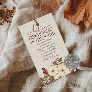 Op de afbeelding: Een rechthoekig, gebroken wit papieren label met touw, voorzien van de tekst Autumn SIMMERING POTPOURRI, instructies om ingrediënten aan een steelpan met 2 kopjes water toe te voegen, en een aquarelillustratie van appelschijfjes en kaneelstokjes.