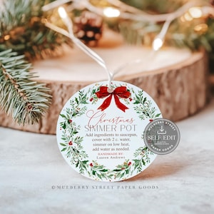 Op de afbeelding: Een rond wit label met een rode strik aan de bovenkant, voorzien van een kransontwerp met groene hulst en bessen. De tekst luidt: Christmas SIMMER POT, Add ingredients to saucepan, cover with 2 c. water, simmer on low heat, add water as needed.