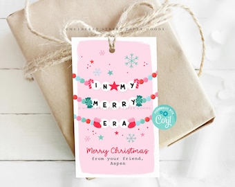 May include: Pink Christmas gift tag with a white border. The tag features a garland of candy canes, snowflakes, and colourful beads with the words "In My Merry Era" in white letters. The text "Merry Christmas from your friend, Aspen" is printed below the garland.