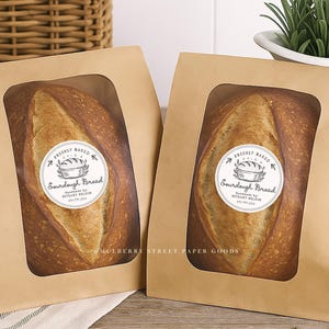 Puede incluir: Dos hogazas de pan de masa madre en bolsas de papel marr&oacute;n con ventanas transparentes. Cada bolsa tiene una pegatina circular que dice "Freshly Baked Sourdough Bread". El pan tiene una corteza dorada.