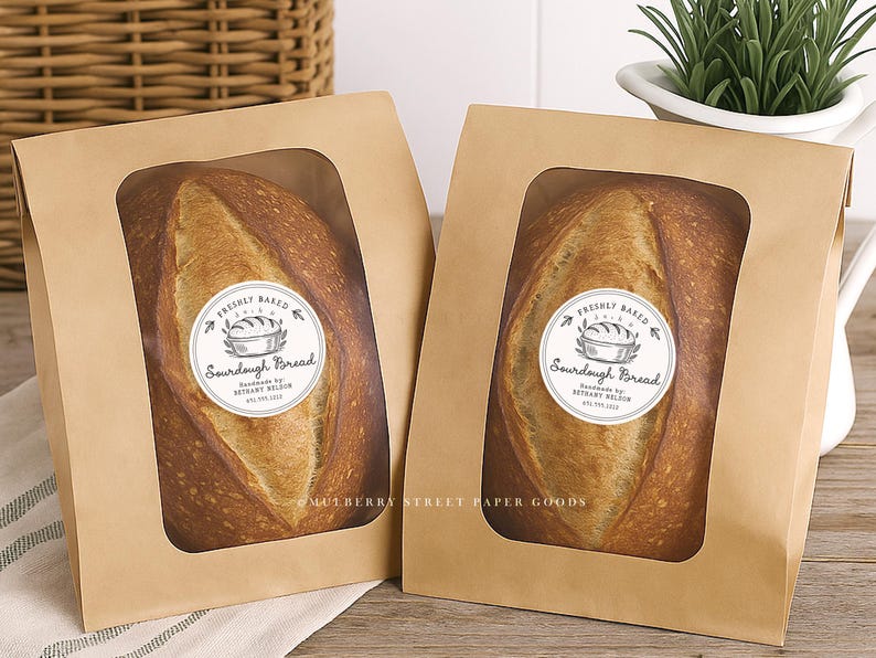 Puede incluir: Dos hogazas de pan de masa madre en bolsas de papel marr&oacute;n con ventanas transparentes. Cada bolsa tiene una pegatina circular que dice "Freshly Baked Sourdough Bread". El pan tiene una corteza dorada.