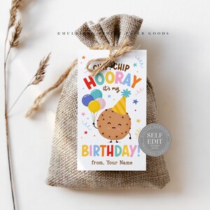 Op de afbeelding: Een kleine jute cadeauzak, vastgebonden met touw, met een verjaardagslabel. Het label leest "Chip Chip Hooray it's my Birthday!" met een cartoon koekjesfiguur met ballonnen. De tag is versierd met kleurrijke sterren en ballonnen.
