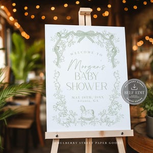 Pu&ograve; includere: Un cartello bianco su un cavalletto con la scritta "Benvenuti al Baby Shower di Morgan" in verde. Il cartello include la data e il luogo ed &egrave; decorato con illustrazioni floreali e fiocchi. Un adesivo con "Self Edit" &egrave; a destra.