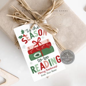 Może przedstawiać: Zapakowany prezent z ozdobną etykietą. Etykieta głosi "'Tis the SEASON to be READING" z ilustracjami książek i płatków śniegu. Prezent jest owinięty w brązowy papier i związany rafią.