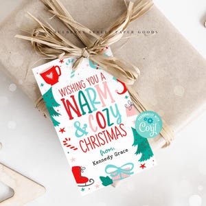 Puede incluir: Una etiqueta de regalo blanca con una ilustración roja, rosa y azul de una taza, bastones de caramelo, un árbol de Navidad y un regalo. El texto dice "Wishing you a warm & cozy Christmas from, Kennedy Grace". Un círculo azul con el texto "PERSONALIZE WITH Coyl!" está en la esquina.