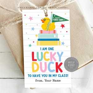 Könnte beinhalten: Ein Geschenkanhänger mit einer gelben Quietscheente mit Brille und einer grünen Flagge mit der Aufschrift "Happy First Day!". Der Anhänger lautet "I am one lucky duck to have you in my class!" mit farbenfrohem Text.
