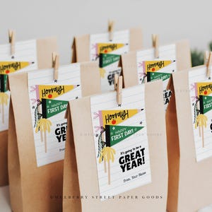 Puede incluir: Varias bolsas de papel marr&oacute;n con etiquetas blancas sujetas con pinzas de madera. Las etiquetas dicen "Hooray! Happy First Day! It's going to be a GREAT YEAR!" con un dise&ntilde;o de bander&iacute;n colorido. Las bolsas son para un evento de regreso a clases.