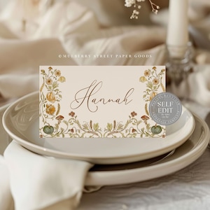 May include: A place card with the name "Hannah" in elegant script, surrounded by floral illustrations in shades of yellow, green, and brown. The card is set on a white plate, suggesting a wedding or event setting.
