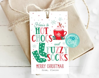 Pode incluir: Uma etiqueta de presente branca com texto vermelho, verde e rosa que diz "Here's to hot chocs and fuzzy socks Merry Christmas from, Claire". A etiqueta também apresenta uma caneca vermelha com um marshmallow branco e um padrão de floco de neve verde e branco. Um padrão de floco de neve verde e branco também está na etiqueta. A etiqueta tem um círculo turquesa com o texto "Personalizar com Cory!" em branco.