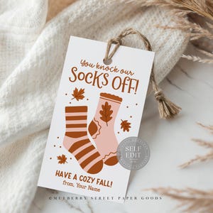 May include: A white tag with the text "You knock our Socks OFF!" and "HAVE A COZY FALL!" features illustrations of socks and fall leaves. The tag is tied with a natural twine.
