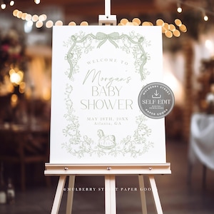 Pu&ograve; includere: Un cartello bianco per baby shower su un cavalletto di legno, con un design a ghirlanda floreale verde. Il cartello recita "Benvenuti al baby shower di Morgan" con la data e il luogo. Un logo circolare con la scritta "Self Edit" &egrave; a destra.