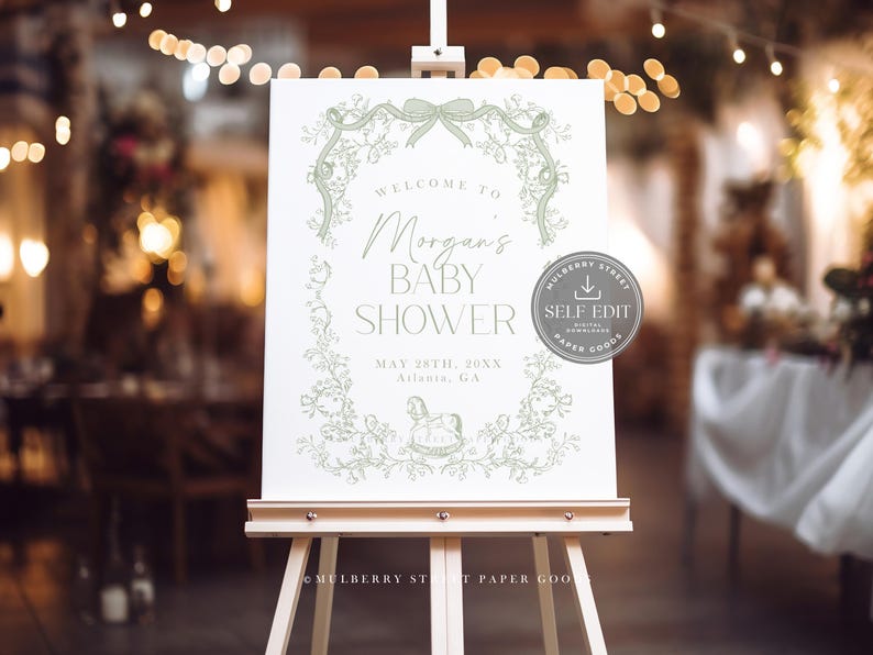 Pu&ograve; includere: Un cartello bianco per baby shower su un cavalletto di legno, con un design a ghirlanda floreale verde. Il cartello recita "Benvenuti al baby shower di Morgan" con la data e il luogo. Un logo circolare con la scritta "Self Edit" &egrave; a destra.