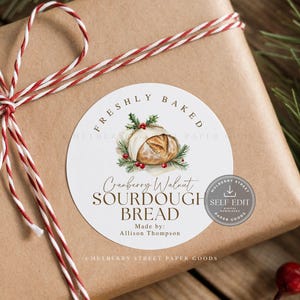 May include: A gift wrapped in brown paper, tied with red and white twine. A round sticker on top reads "Freshly Baked Cranberry Walnut Sourdough Bread." The sticker features a watercolor illustration of bread and greenery.