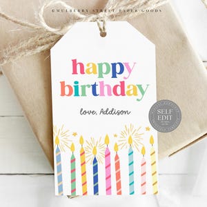 Puede incluir: Una etiqueta de regalo de cumpleaños blanca con las palabras "feliz cumpleaños" en letras coloridas. La etiqueta tiene un diseño de velas en la parte inferior y está atada a un regalo envuelto con hilo.