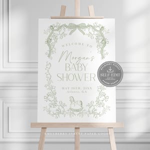 Pu&ograve; includere: Un cartello per baby shower su un cavalletto di legno. Il cartello presenta il testo "Welcome to Morgan's Baby Shower" in una scrittura elegante, con la data e il luogo sottostanti. Il design include elementi floreali e fiocchi verdi.