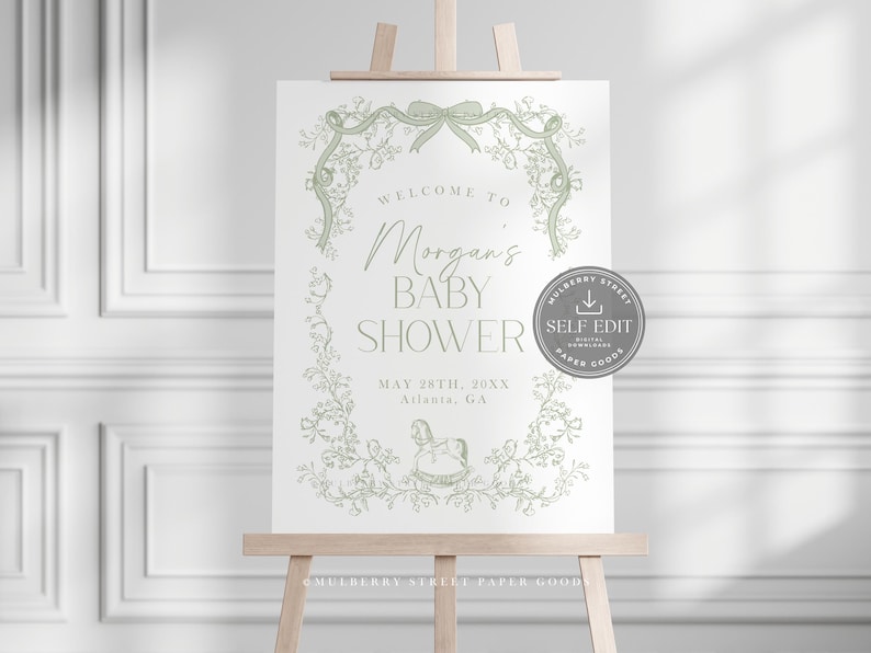 Pu&ograve; includere: Un cartello per baby shower su un cavalletto di legno. Il cartello presenta il testo "Welcome to Morgan's Baby Shower" in una scrittura elegante, con la data e il luogo sottostanti. Il design include elementi floreali e fiocchi verdi.