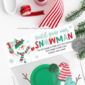 May include: A DIY snowman kit with a green lid, snowman accessories, and a card that reads "Build your own SNOWMAN." The card features a snowman illustration and instructions. Red and white striped ribbon and a sprig of evergreen are in the background.